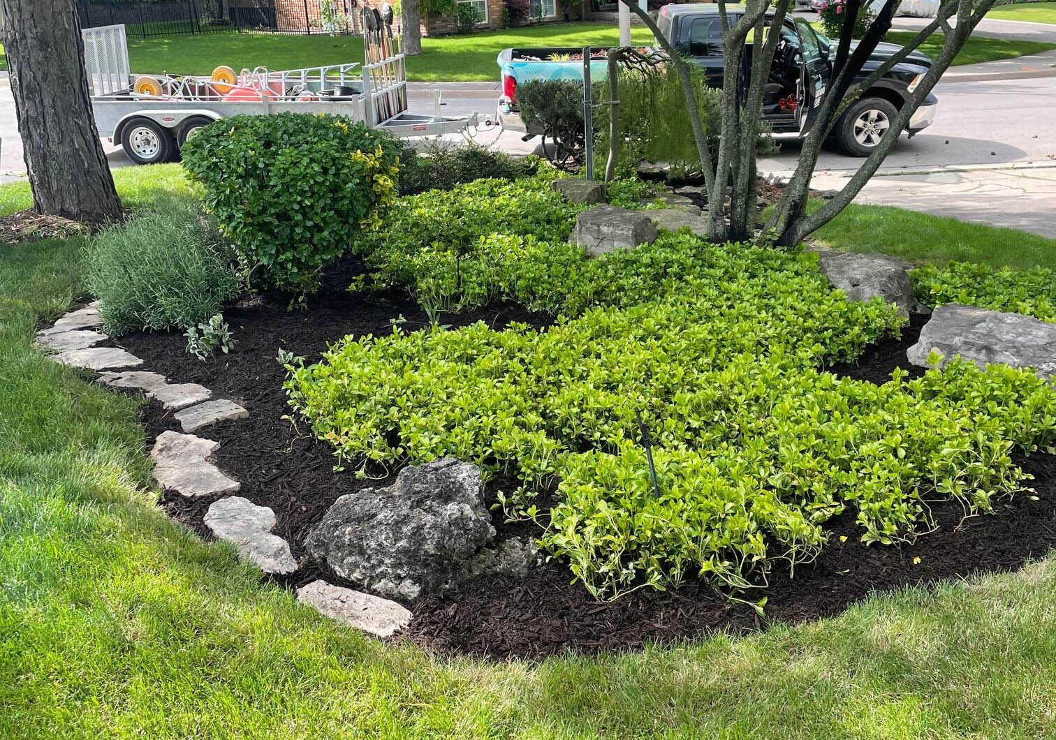 A well-maintained garden with neatly arranged plants and stones sits by a suburban street, where a truck and trailer are parked.
