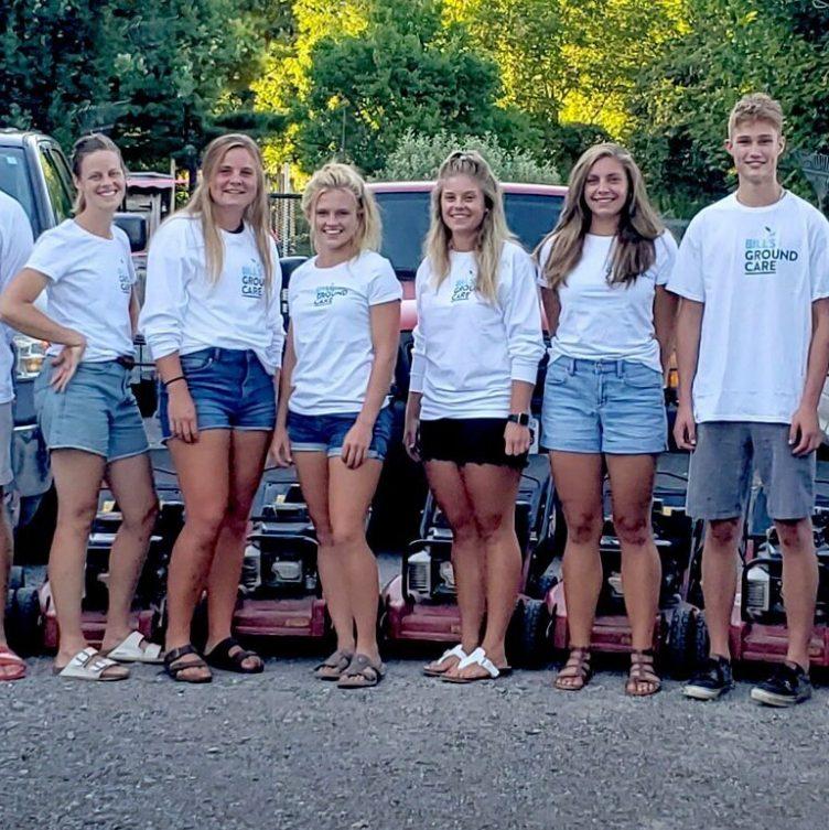A group of nine people wearing "Bill’s Ground Care" shirts, standing outdoors in front of lawnmowers, with trees in the background.