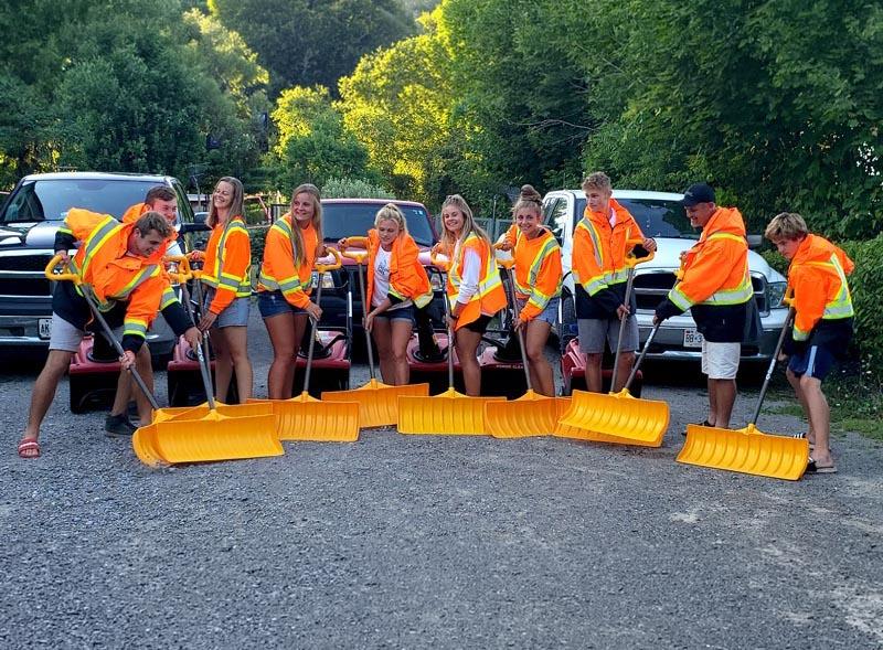 A group of people in fluorescent jackets poses with orange snow shovels in front of trucks, standing on a gravel surface outdoors.