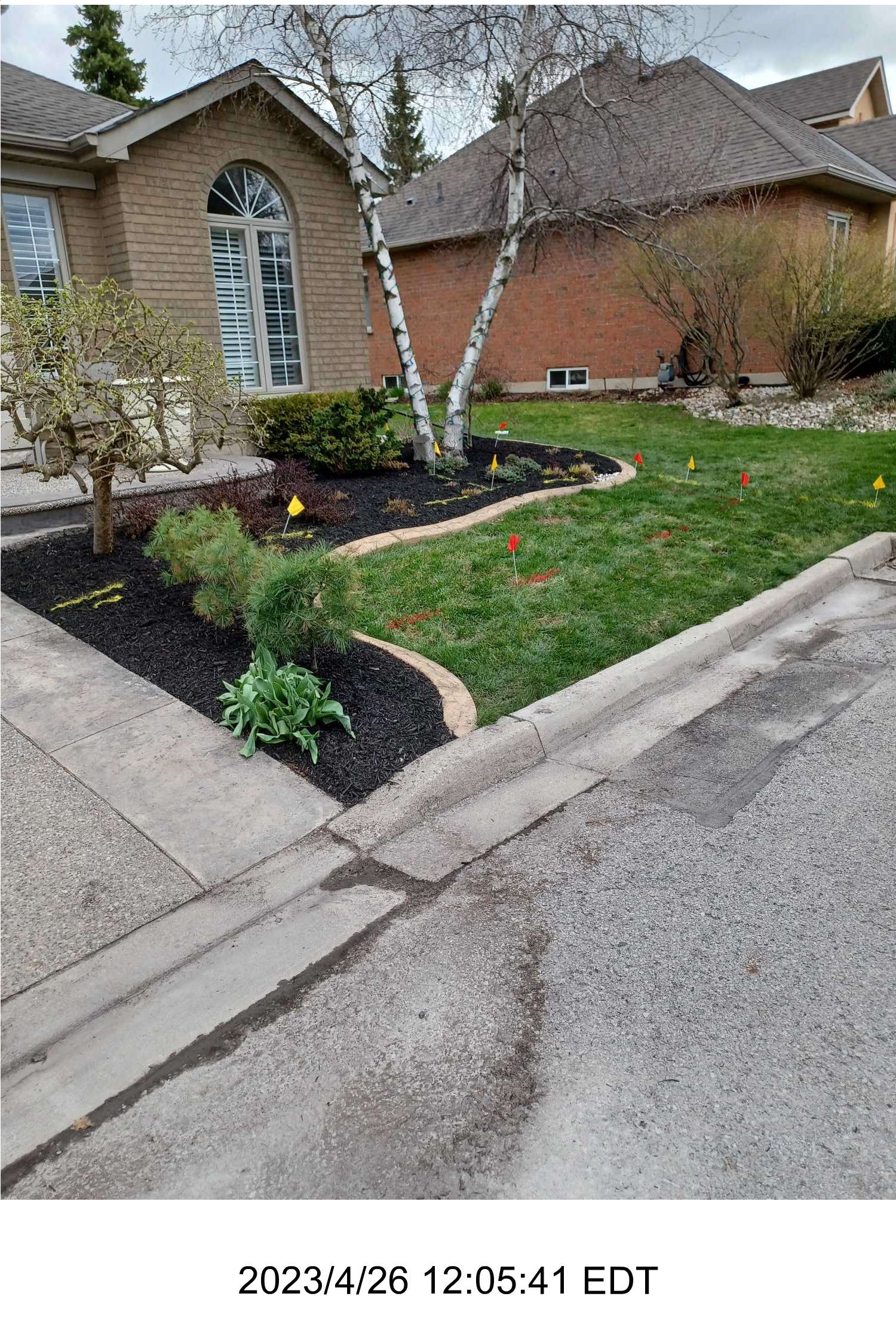 A suburban house with manicured garden, featuring mulch, small trees, and colorful markers on the lawn. No recognizable landmarks or historical buildings.