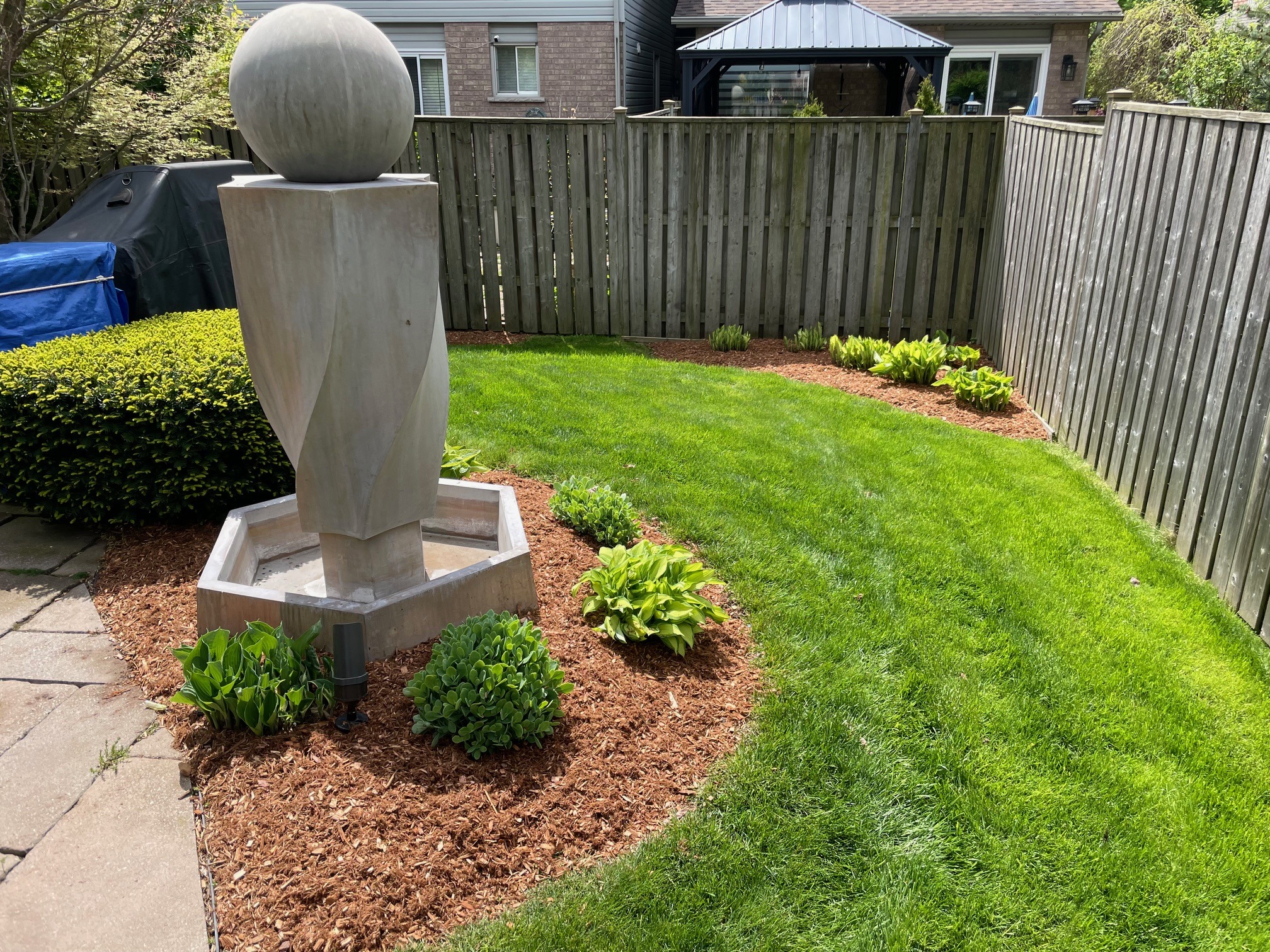 A neatly landscaped garden features a modern sculpture, surrounded by mulch and young plants, enclosed by a wooden fence and patio area.