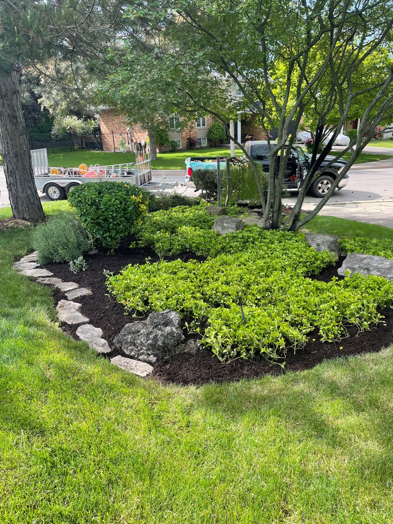A well-maintained garden with neatly arranged plants and stones sits by a suburban street, where a truck and trailer are parked.