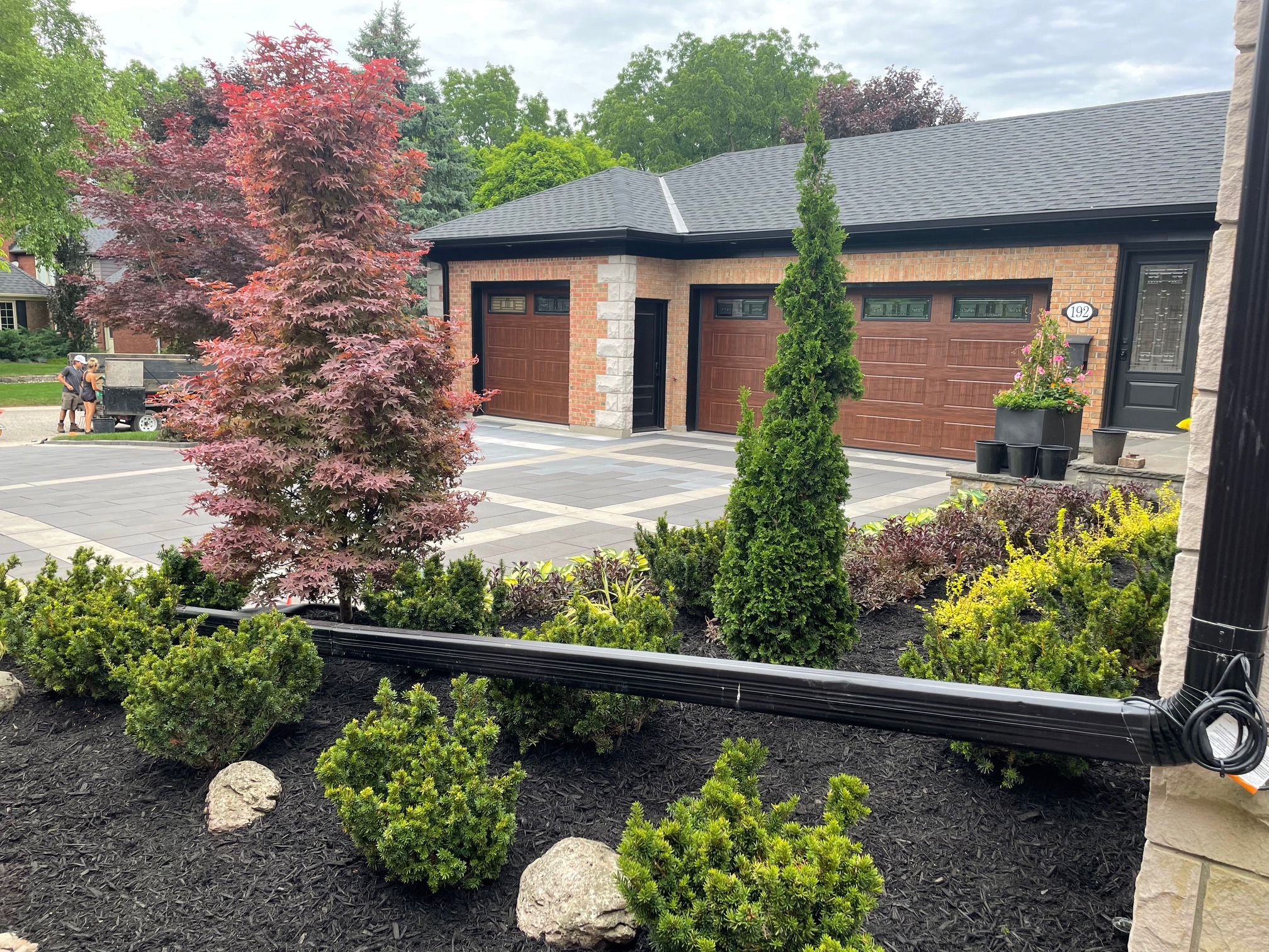 A well-maintained garden with colorful shrubs and trees in front of a modern house. Two people stand near the driveway, talking.
