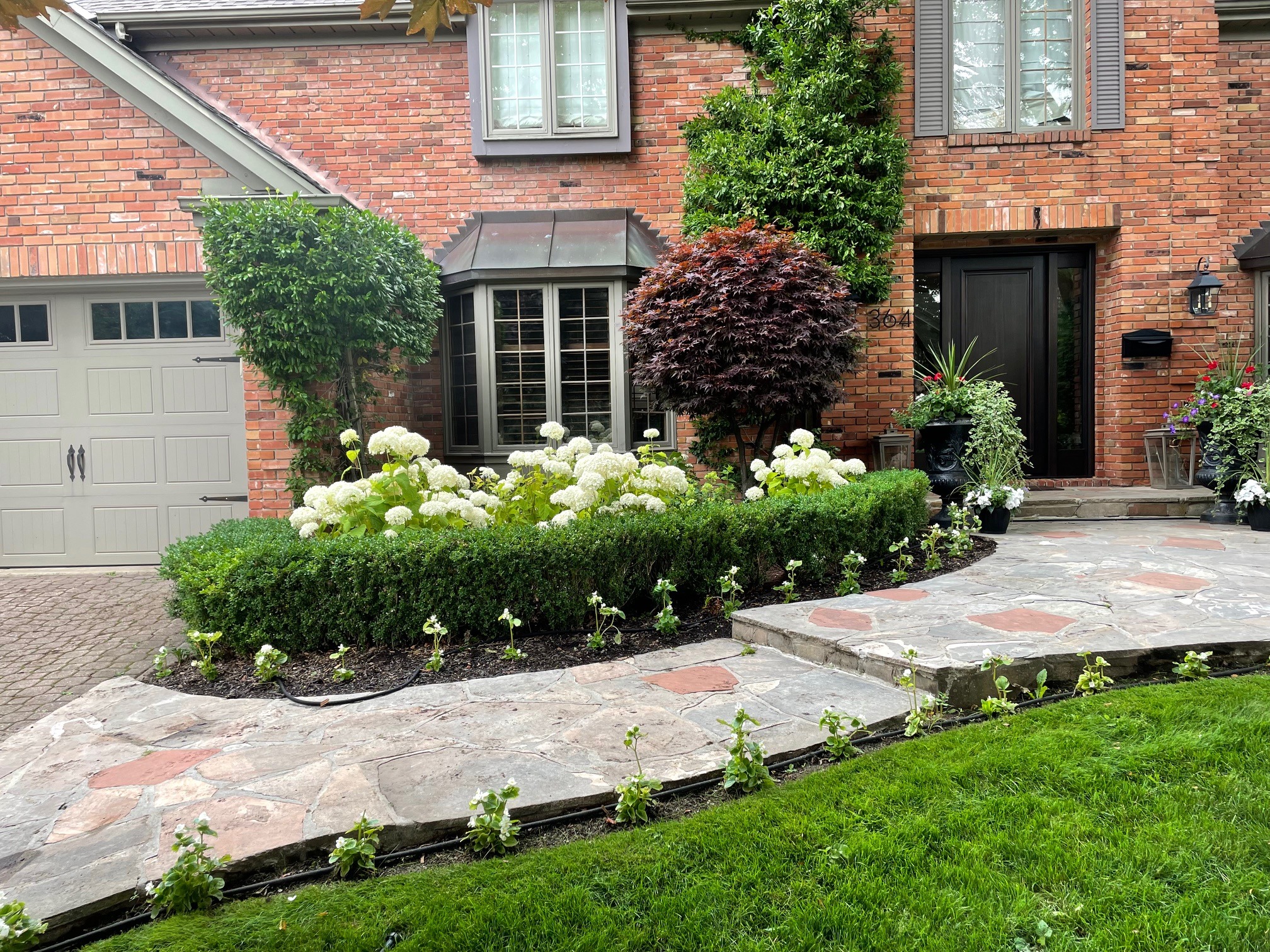 Brick house with well-manicured garden, stone pathway, and various potted plants. Features a two-car garage and a bay window.