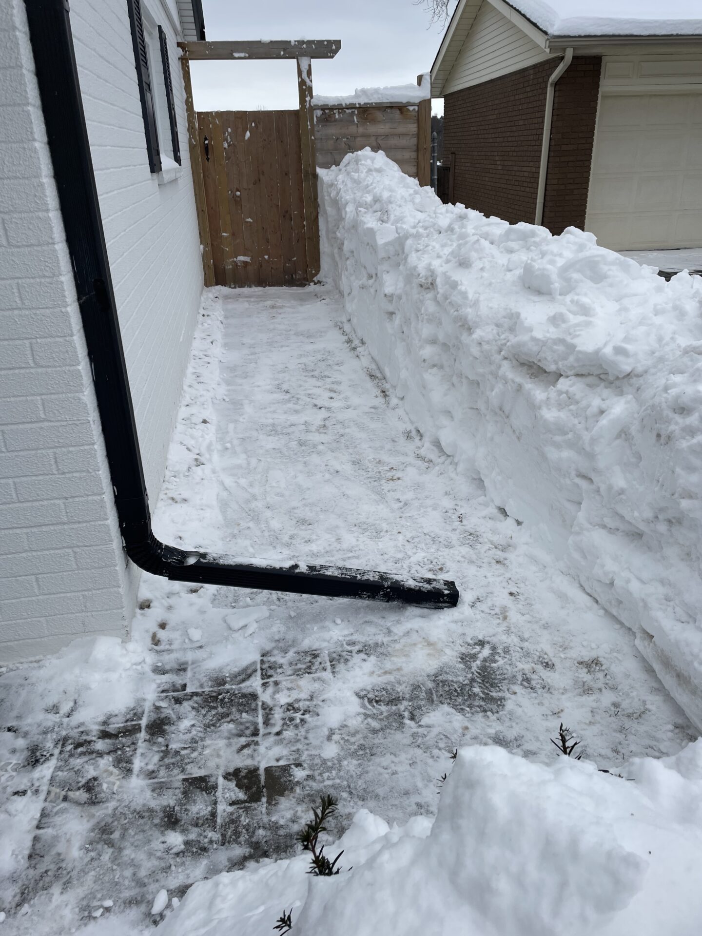 Narrow pathway between two houses with high snow piles. Snow-covered ground drains into a black gutter pipe attached to the wall.