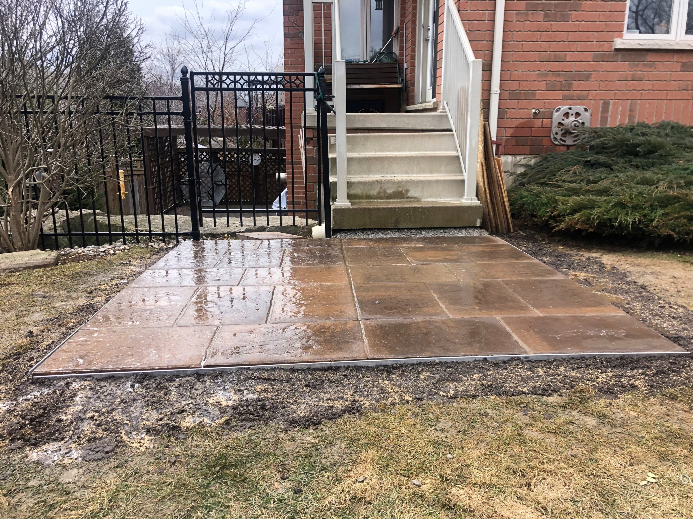 Wet stone patio leads up to brick house with stairs. Black gate, bare trees, and green bushes surround the area.