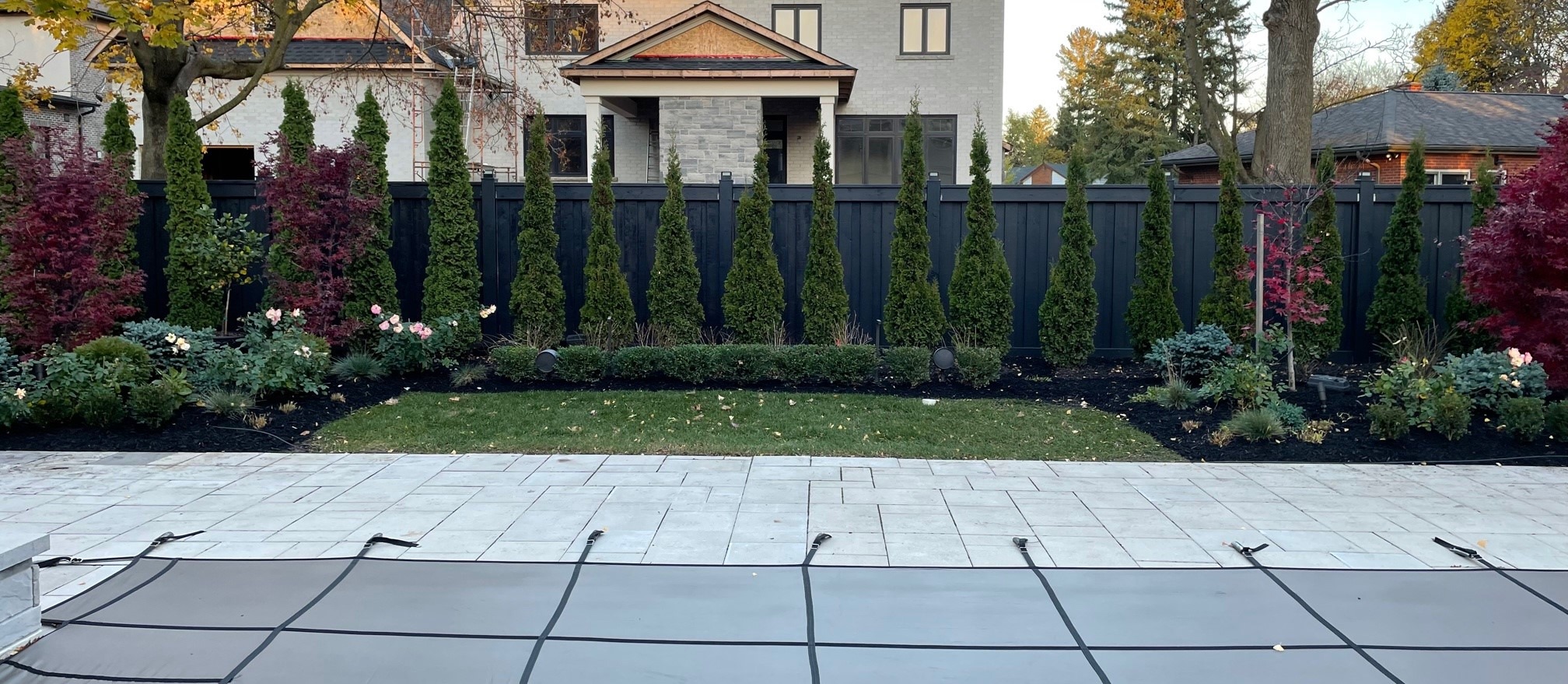 A modern backyard features a covered pool, stone patio, manicured lawn, tall evergreen trees, vibrant shrubs, and a gray house in the background.