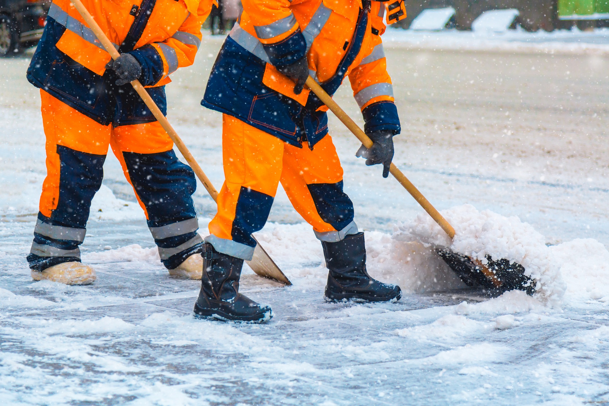 Two persons in orange uniforms shoveling snow on a snowy street, wearing boots and gloves, amidst a wintery atmosphere, focused on removing snow.