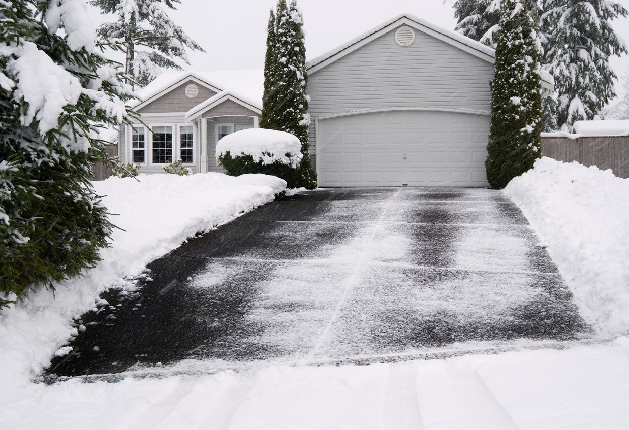 A snow-covered suburban house with a clear driveway sits amid tall trees and neatly trimmed shrubs under an overcast winter sky.