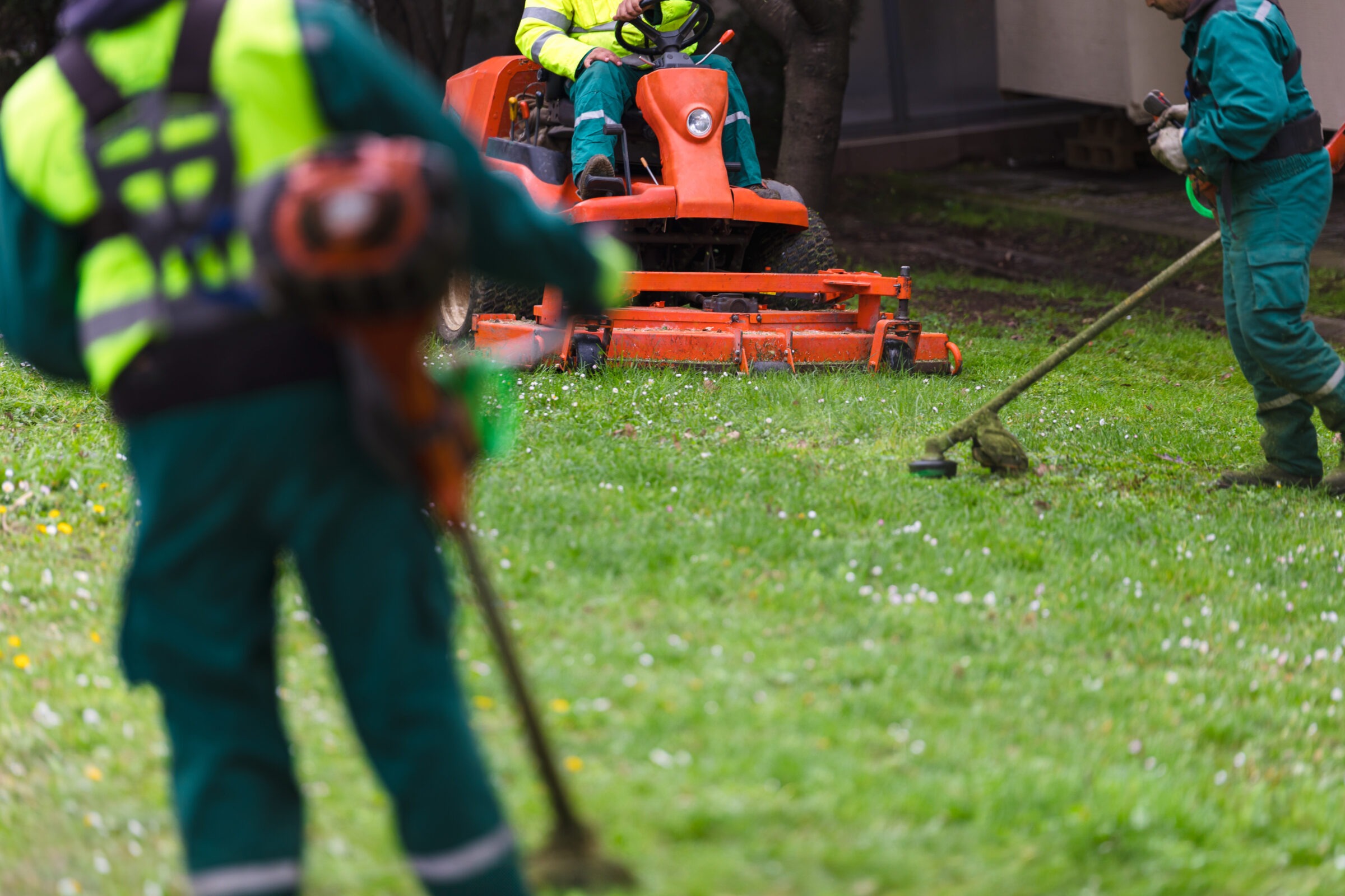 Three persons in reflective clothing maintain a green lawn using a ride-on mower and string trimmers, focusing on the grass around them.