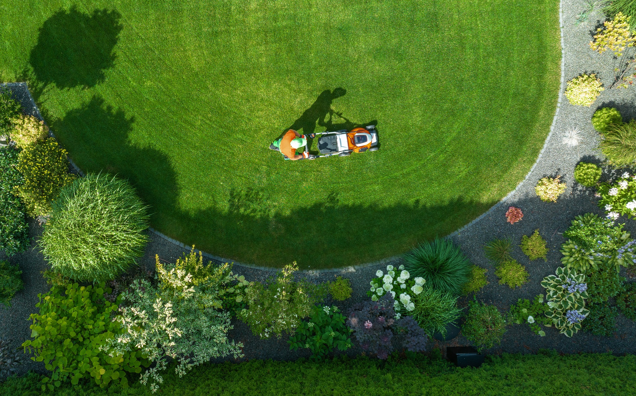 A person in orange mows a circular lawn, surrounded by colorful garden plants and shadows, viewed from above.