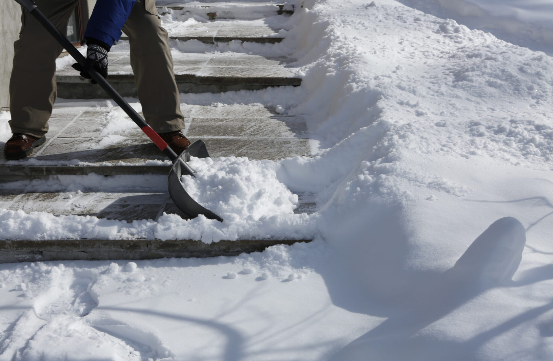 A person shovels snow from a set of concrete steps, surrounded by a thick, undisturbed snow blanket on a sunny winter day.