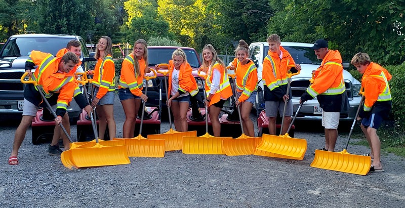 A group of people in fluorescent jackets poses with orange snow shovels in front of trucks, standing on a gravel surface outdoors.